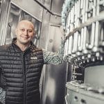 This image shows Adam Broz, brew master standing in front of brewing equipment